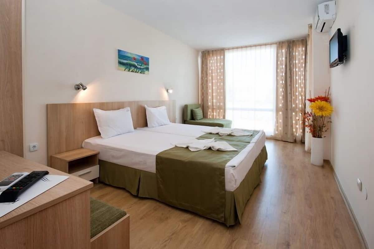 Una habitación de hotel con cama, mesita de noche, un escritorio con silla, un pequeño sofá, televisor montado en la pared y ventana con cortinas.