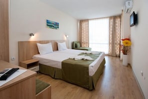 A hotel room with a bed, bedside table, a desk with a chair, a small sofa, a TV mounted on the wall, and a window with curtains.