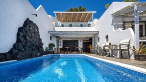 A swimming pool with a stone wall on one side and a white building with a covered patio area on the other. - Plastiras Rooms (Santorini)