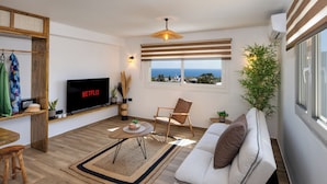 A modern living room with a flat-screen TV, a sofa, a coffee table, and a view of the sea. - Plastiras Rooms (Santorini)