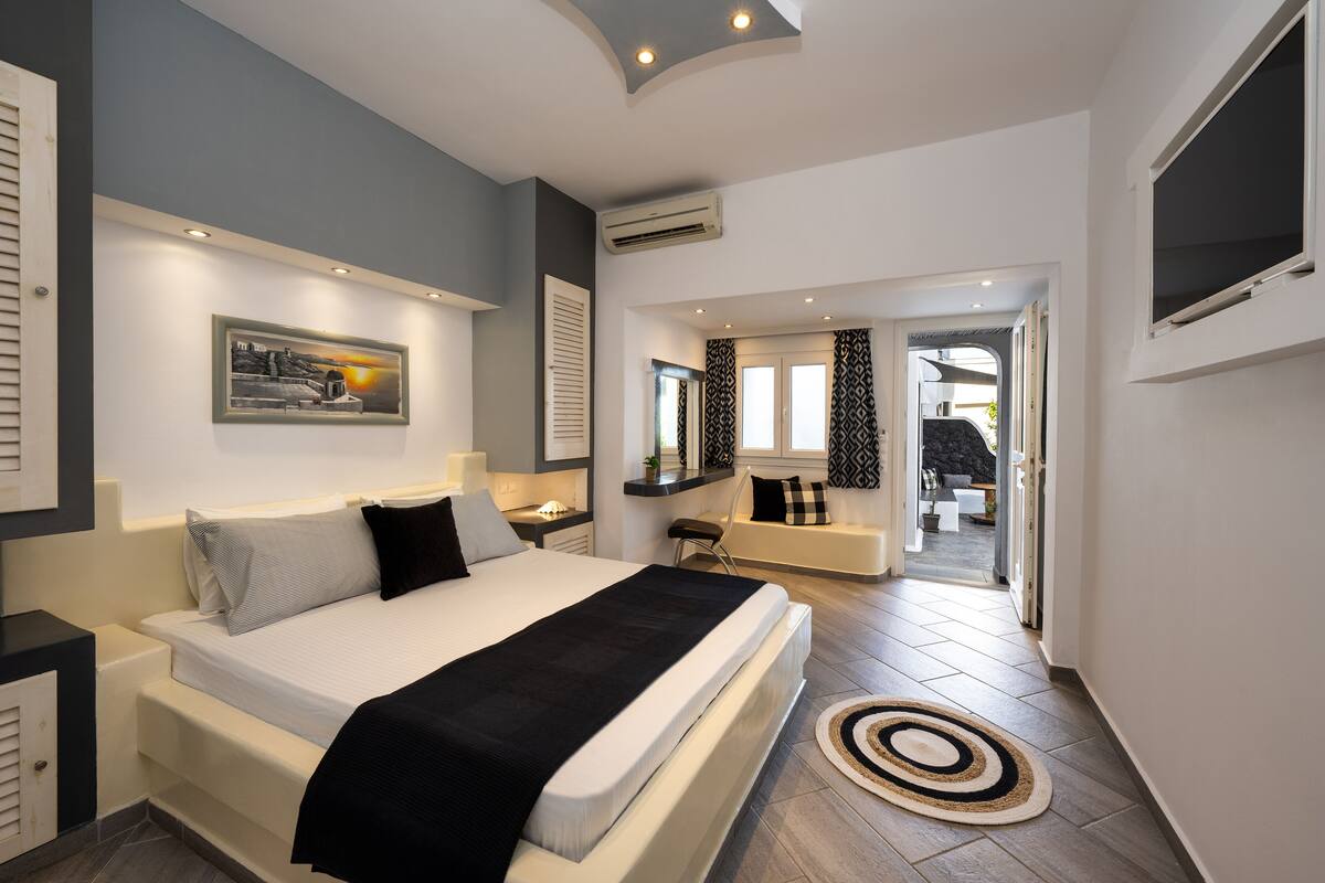 A modern bedroom with a large bed, bedside tables, a nightstand, a window with patterned curtains, and a view into the living area.