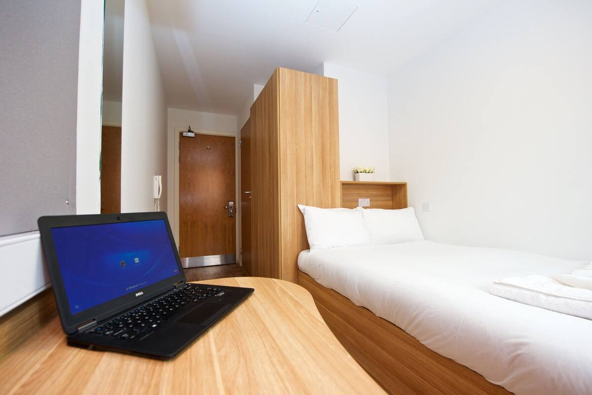 A hotel room with a wooden desk, a laptop, a bed, and a small shelf with a plant.