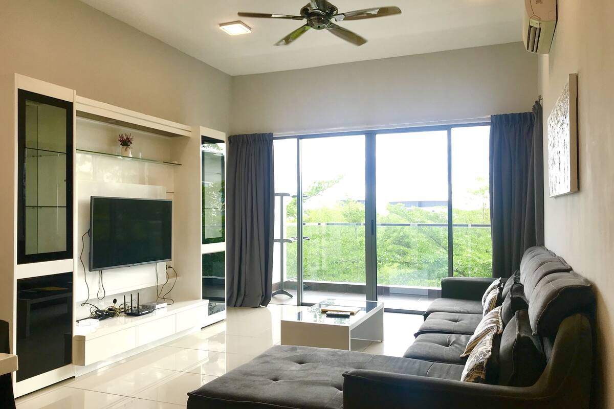 A modern living room with a flat-screen TV, a large window with curtains, a ceiling fan, and a sectional sofa.