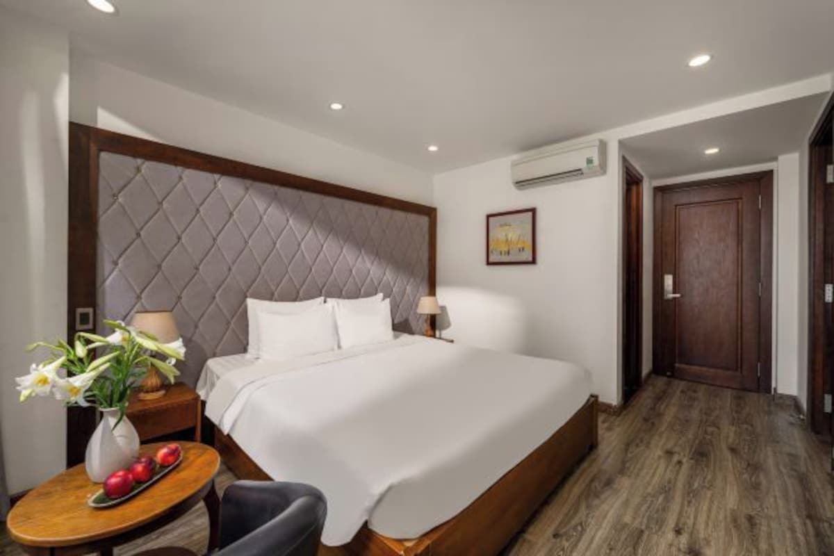 A hotel room with a bed, bedside table with a vase and fruit, a chair, a wooden floor, and a door.