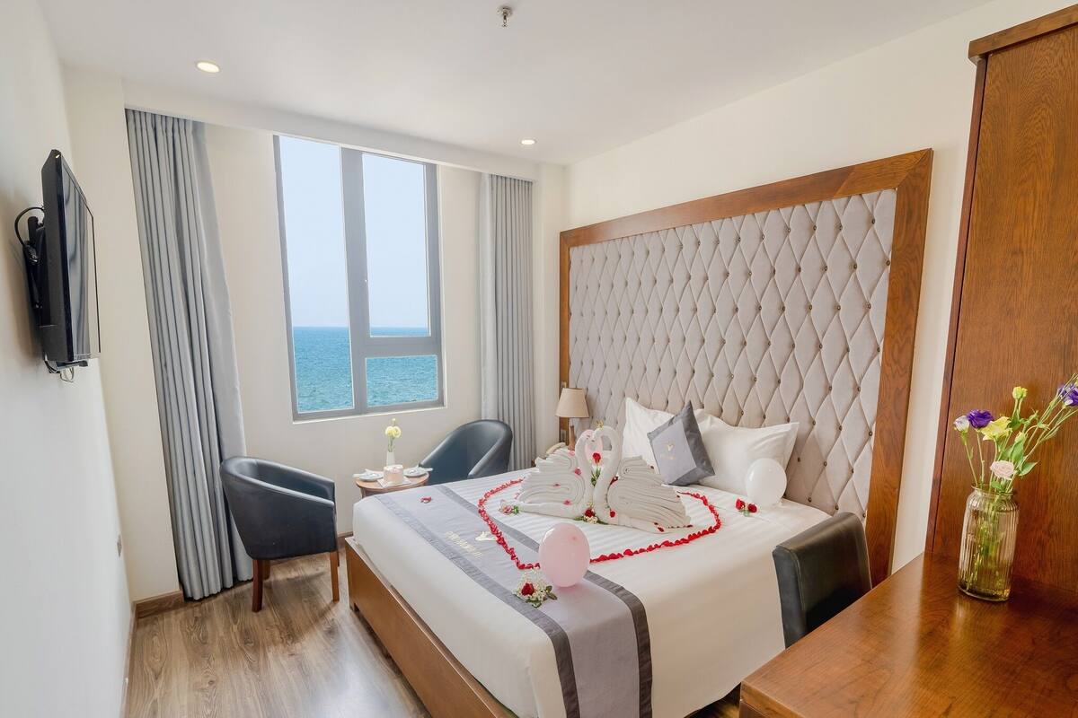 A hotel room with a large bed, a TV, a desk, and a view of the sea.