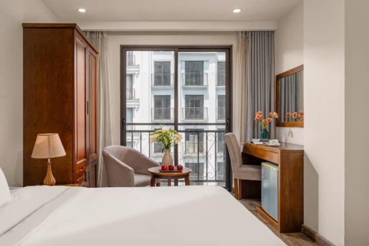 A hotel room with a bed, a desk, a chair, a lamp, a small table with flowers, and a view of buildings through the window.