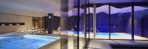 A modern indoor swimming pool with glass walls, a lounge area with chairs, and a visible clock. - Spa Hotel Amsee (Waren (Müritz))