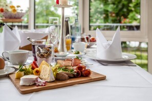 A breakfast buffet with various cheeses, meats, and fruits on a wooden board, set on a white tablecloth with a view of greenery outside. - Spa Hotel Amsee (Waren (Müritz))