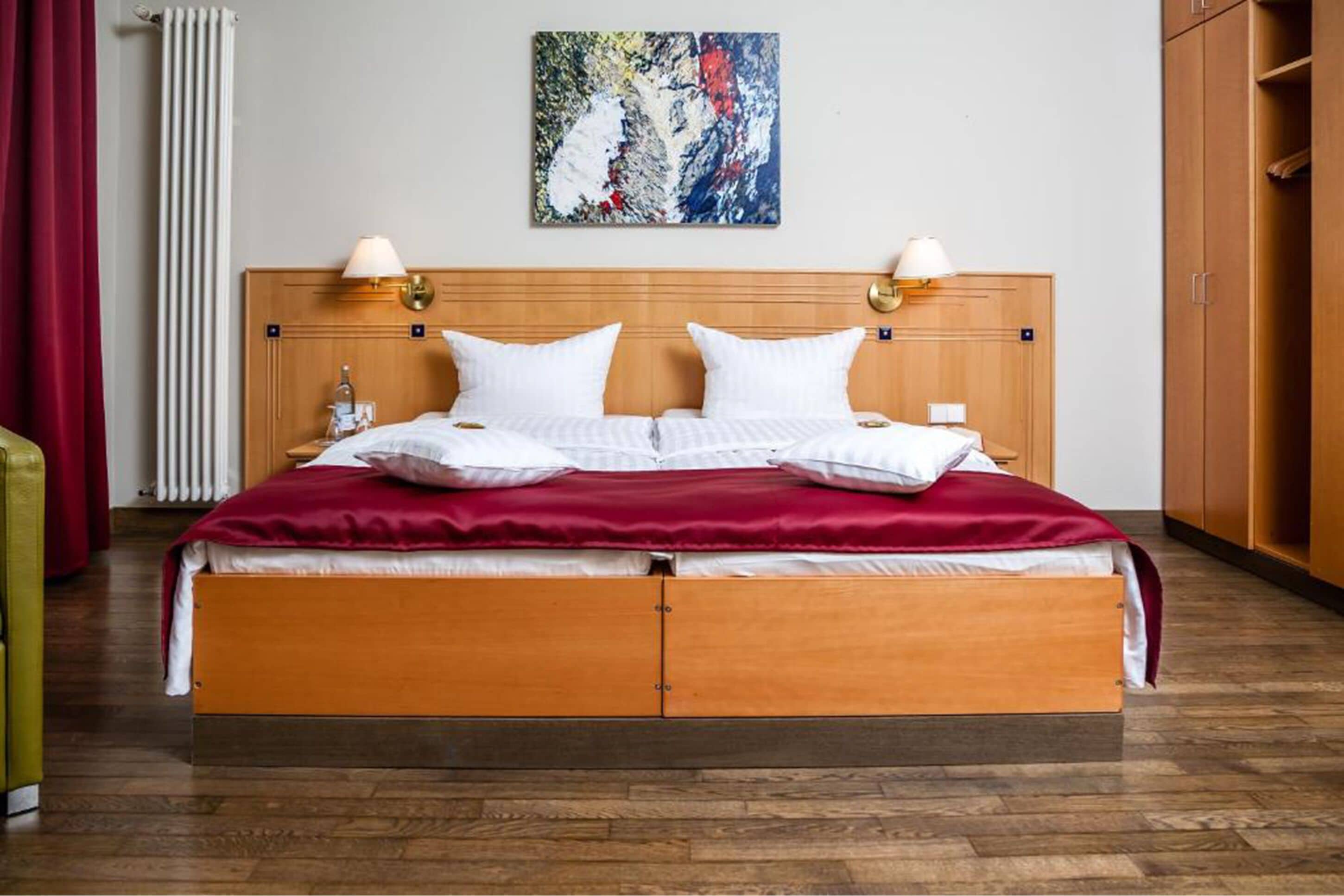 A hotel room with a wooden bed, two bedside lamps, a red blanket, and a painting on the wall.