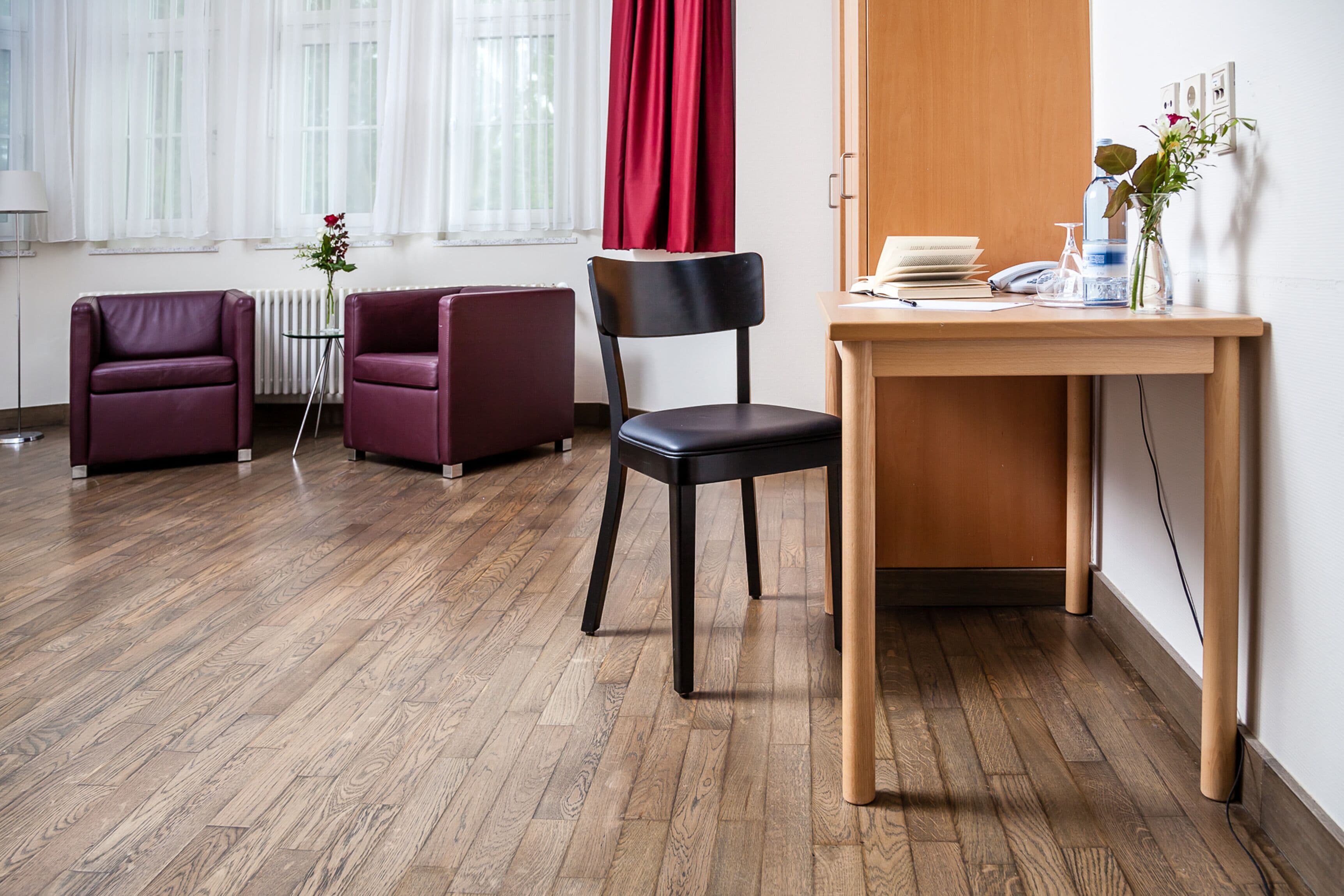 Una habitación con piso de madera, una silla negra, dos sillones morados, un escritorio de madera con un teléfono, un florero con una flor y un radiador.