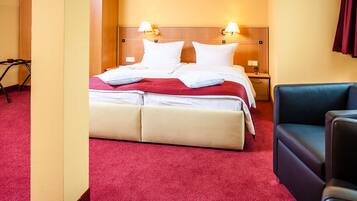 Quarto de hotel com cama, duas luminárias de cabeceira, mesa de cabeceira, poltrona azul e tapete vermelho.