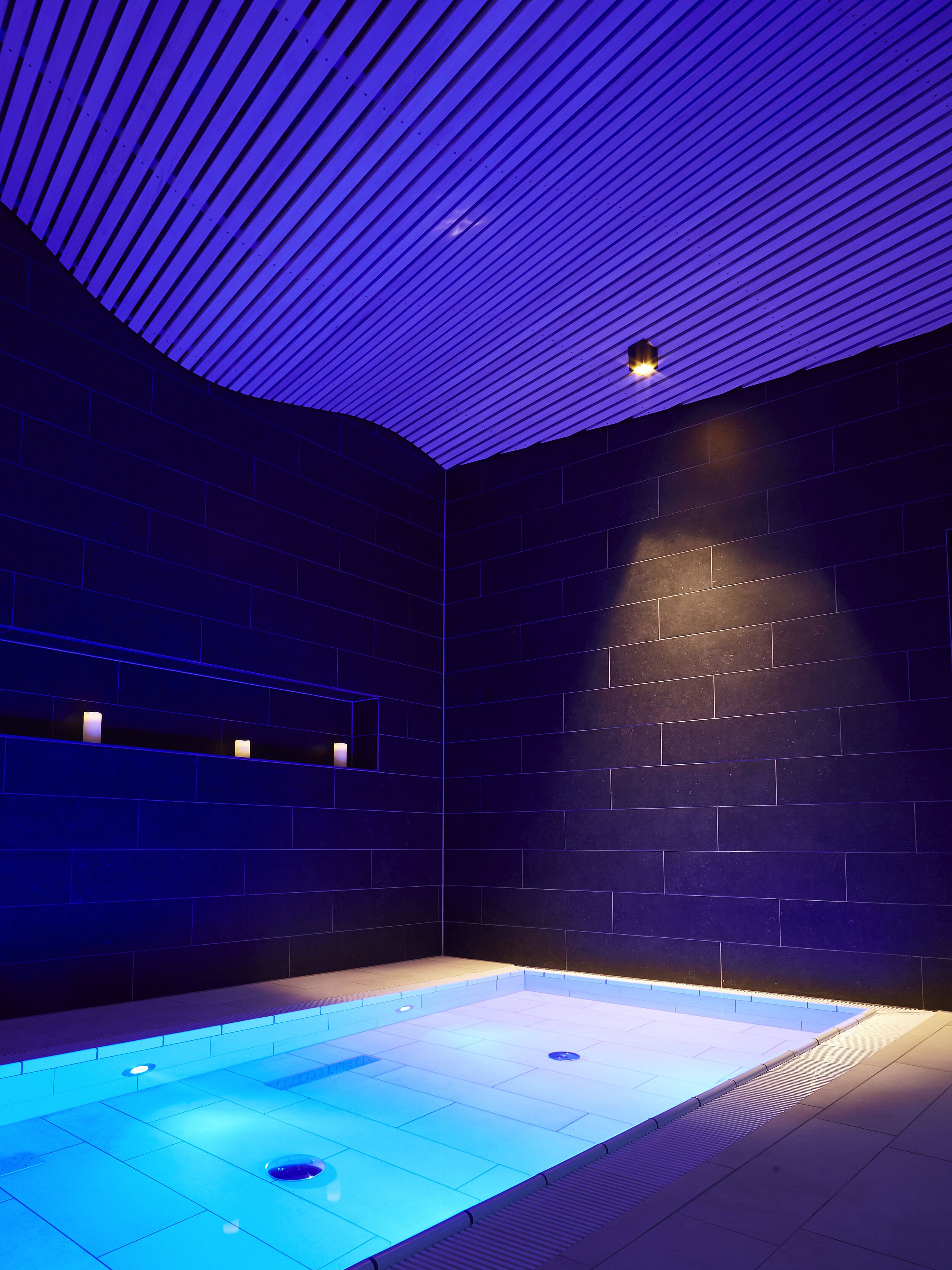 A modern indoor swimming pool with a blue-lit ceiling and a tiled floor.