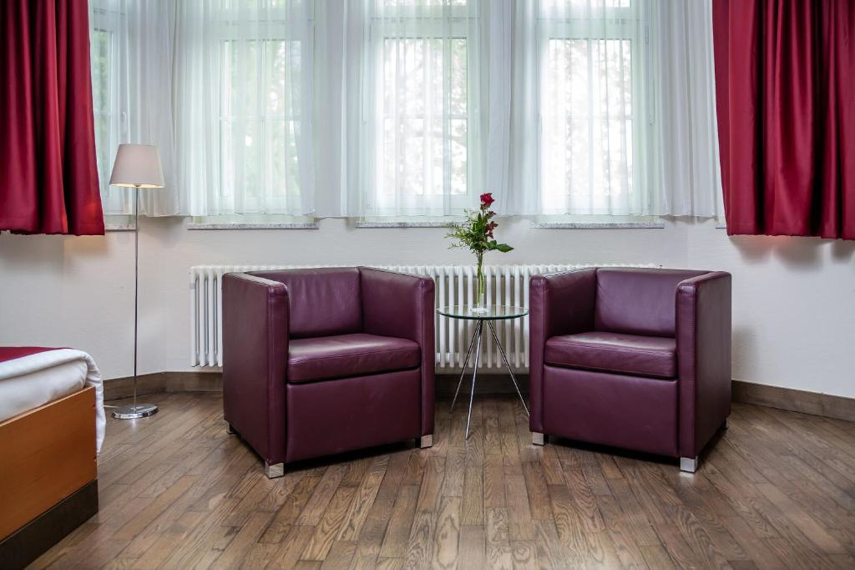 Um quarto com duas poltronas de couro roxo, uma mesa redonda pequena com um vaso de flores, um abajur e um radiador.