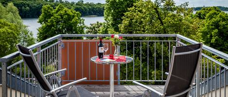 A balcony with two chairs and a small table, overlooking a lake with trees and a wooden deck.