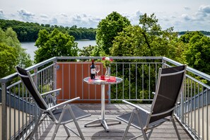 A balcony with two chairs and a small table, overlooking a lake with trees and a wooden deck. - Spa Hotel Amsee (Waren (Müritz))