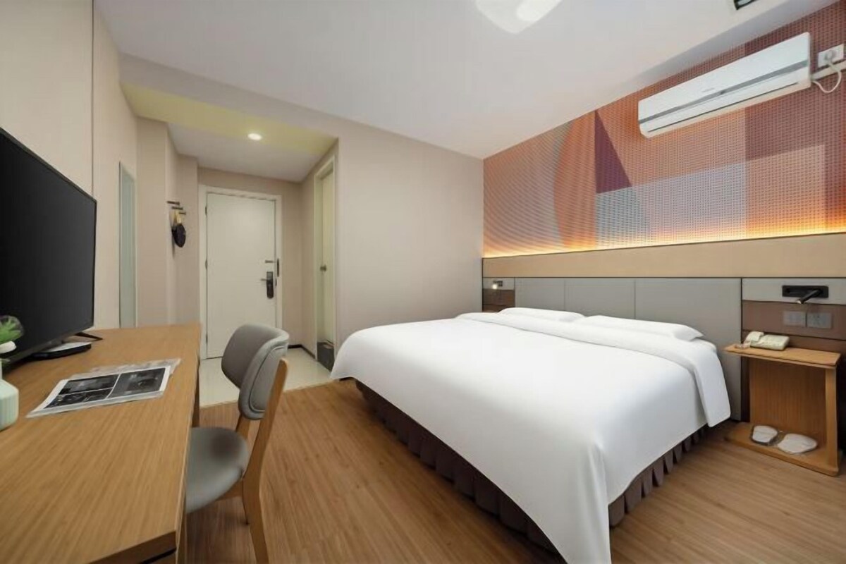 A hotel room with a bed, a desk, a chair, a TV, and a wall-mounted air conditioner.