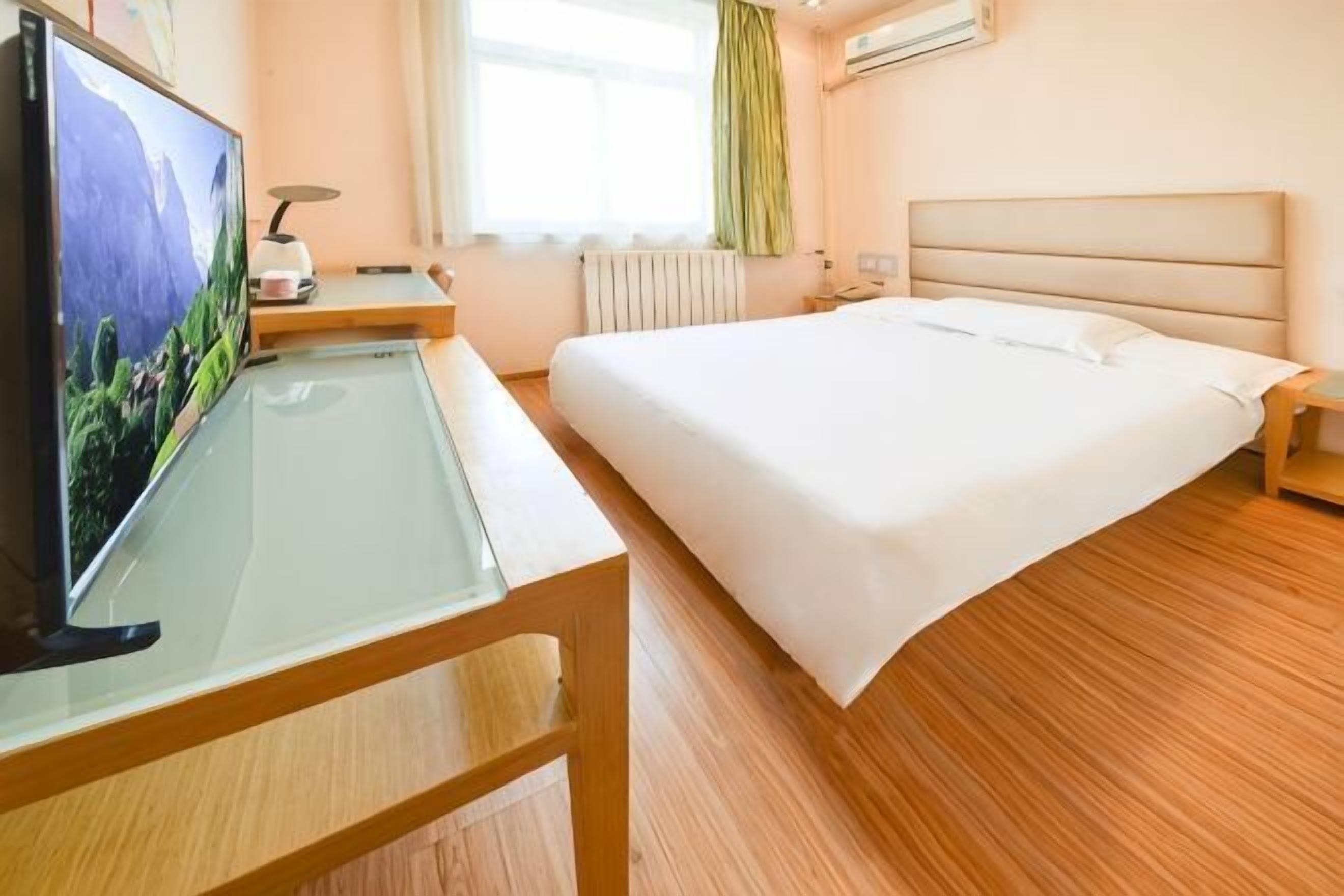 A hotel room with a bed, a TV on a glass table, a bedside table, and a window with curtains.