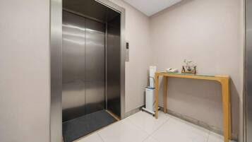 Un ascensor con detalles metálicos, una pequeña mesa con un teléfono y elementos decorativos, y un piso de baldosas.