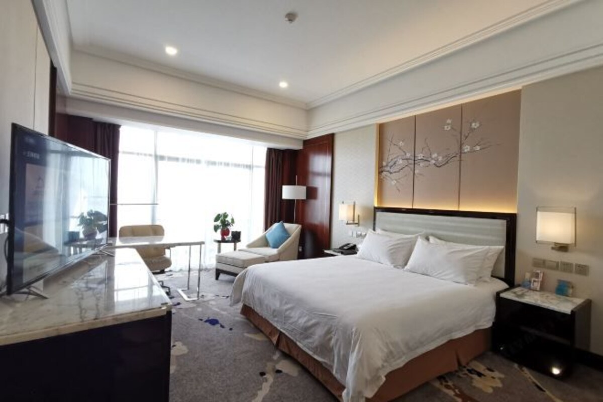 A hotel room with a large bed, a desk with a flat-screen TV, a chair, a nightstand, and a wall-mounted lamp.