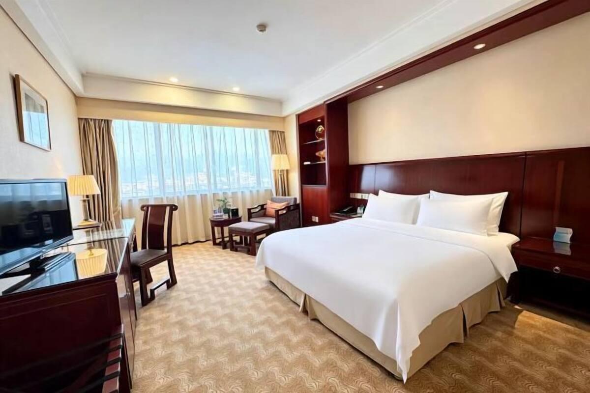 Ein Hotelzimmer mit einem großen Bett, einem Schreibtisch mit Fernseher, einem Sessel, einem kleinen Tisch und einem Fenster mit Vorhängen.