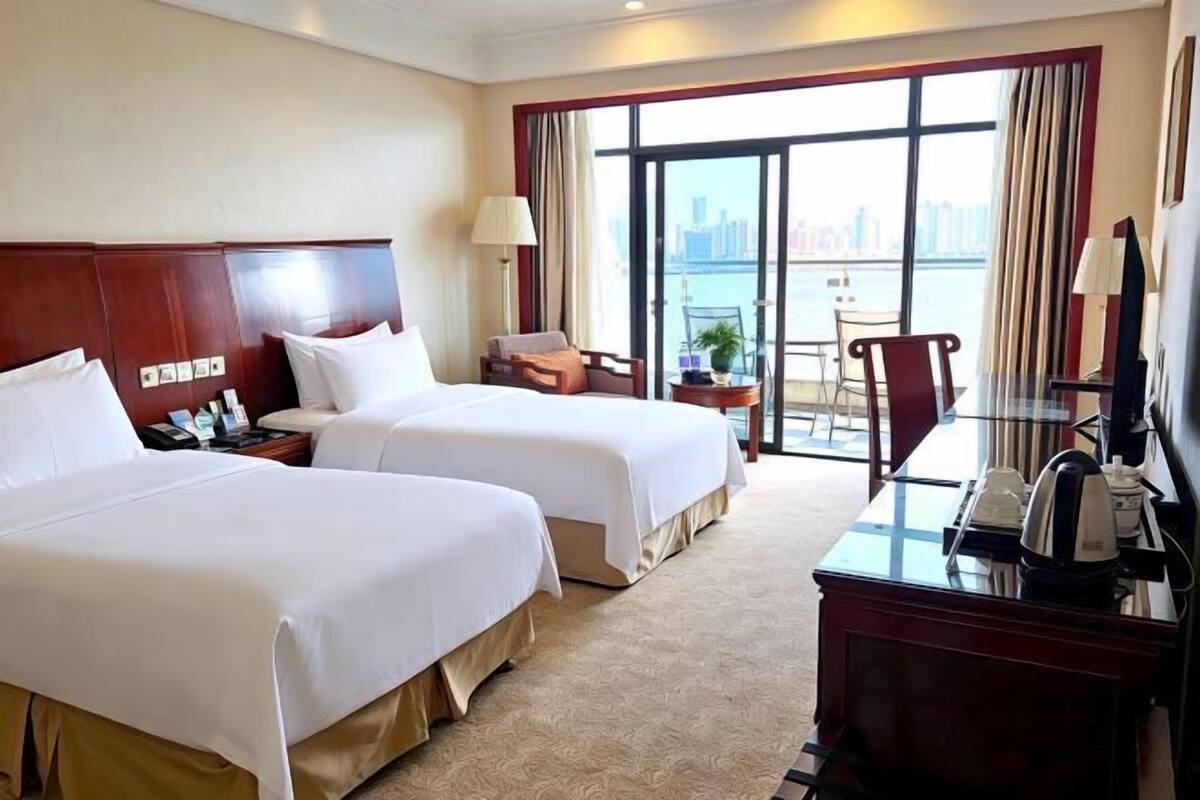 Ein Hotelzimmer mit zwei Betten, einem Schreibtisch, einem Sessel und einer Lampe, mit Blick auf eine Stadtlandschaft durch große Fenster.