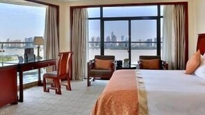 A hotel room with a large bed, a desk, and a chair. There is a view of a cityscape through the windows. - Mengjiang Hotel (Wenzhou)