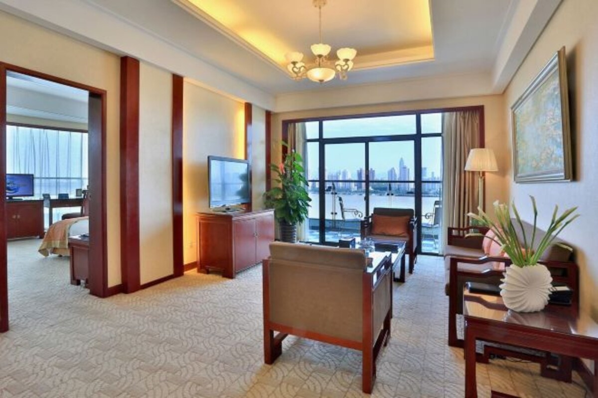 A hotel room with a large window offering a city view, a seating area with a sofa and chairs, a television, and a desk with a plant.