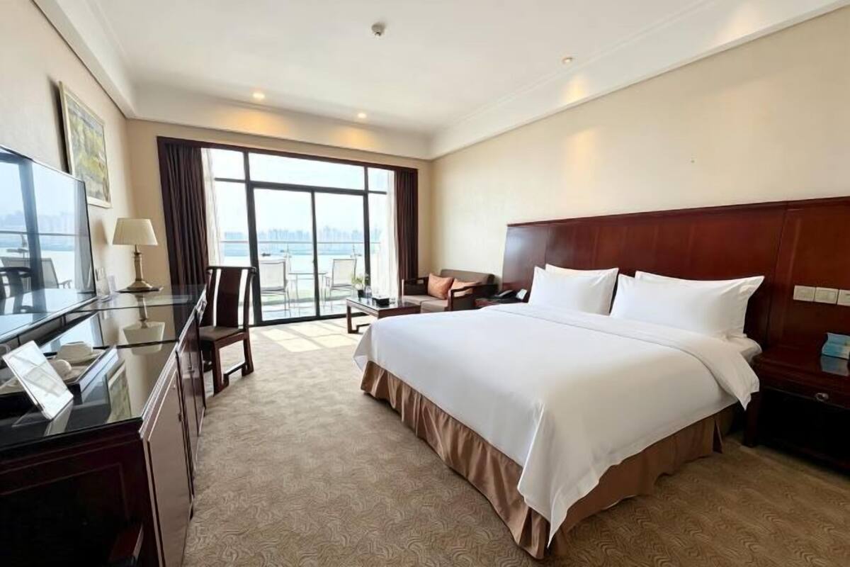 Ein Hotelzimmer mit einem großen Bett, einem Schreibtisch mit Lampe, einem Sessel und Blick auf die Stadt durch große Fenster.