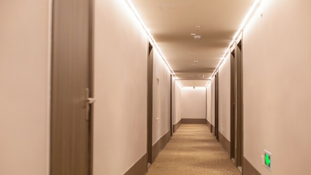 A long, narrow hotel corridor with doors on both sides, carpeted floors, and overhead lighting.