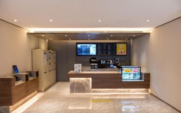 Zona de recepción de un hotel con una pantalla digital, una máquina de café y un enfriador de bebidas.