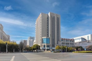 A modern building with a central glass facade, surrounded by a wide street and other buildings. - Fuyuan Hotel (Zhengzhou East Station Provincial Government Branch) (Zhengzhou)