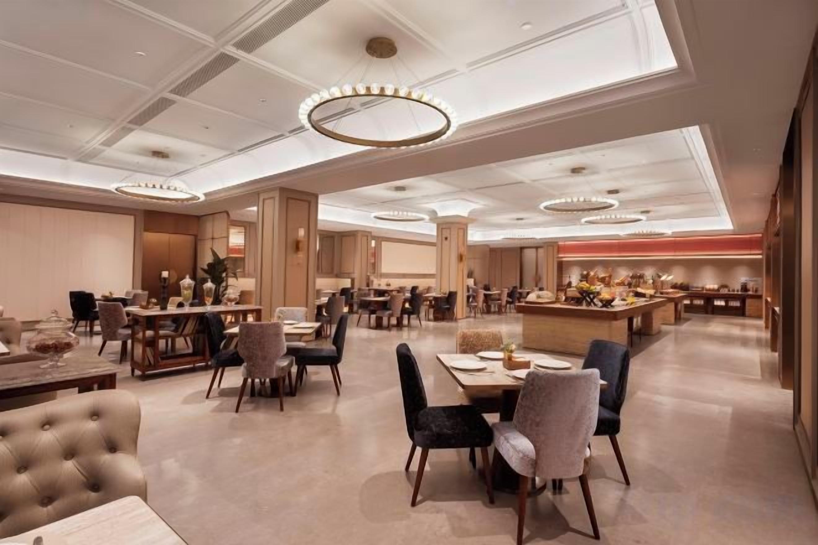 A spacious hotel dining area with multiple tables and chairs, a central bar area, and a high ceiling with modern lighting fixtures.