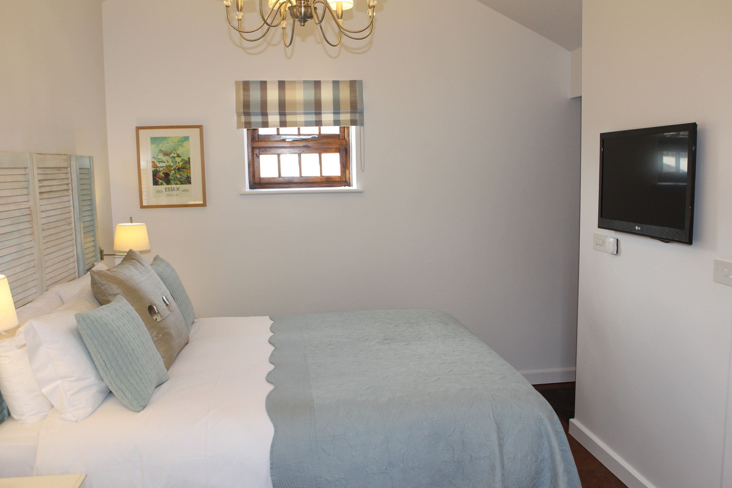Un dormitorio con cama, televisor, ventana con persianas, candelabro y un cuadro enmarcado en la pared.