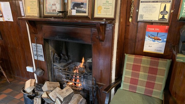 A traditional fireplace with logs, a chair, and various framed certificates and notices on the wall. - The Compasses Inn (Chelmsford)
