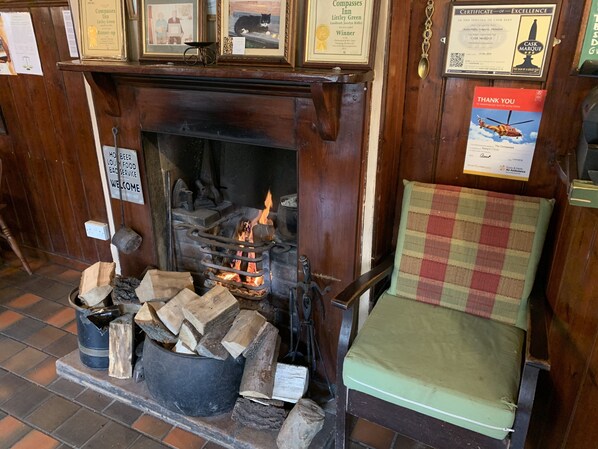 A traditional fireplace with logs, a chair, and various framed certificates and notices on the wall. - The Compasses Inn (Chelmsford)