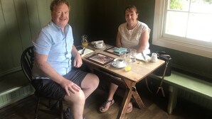 Two people seated at a wooden table with a green cushioned bench behind them. The table has various items including a pitcher, cups, and a plate with a picture. - The Compasses Inn (Chelmsford)
