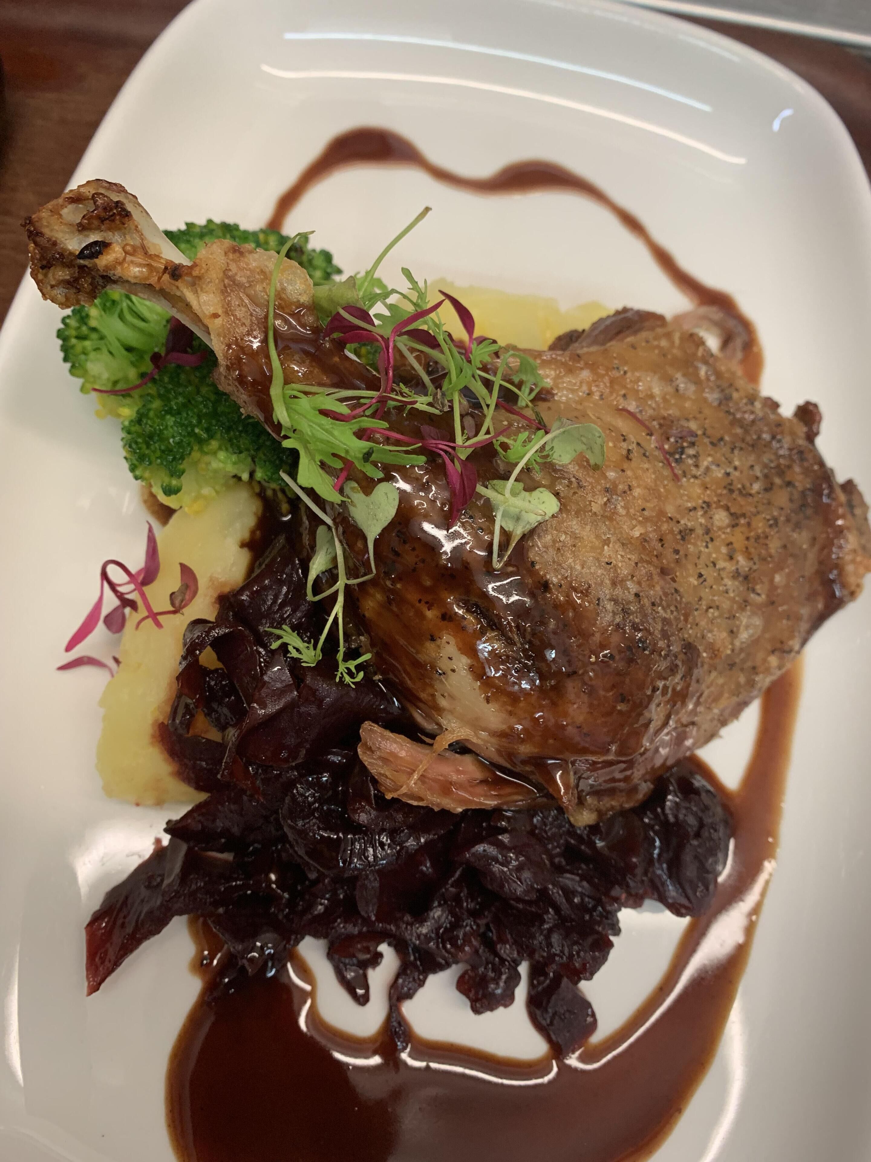 A plate with a roasted duck leg, broccoli, purple cabbage, and a dark sauce.
