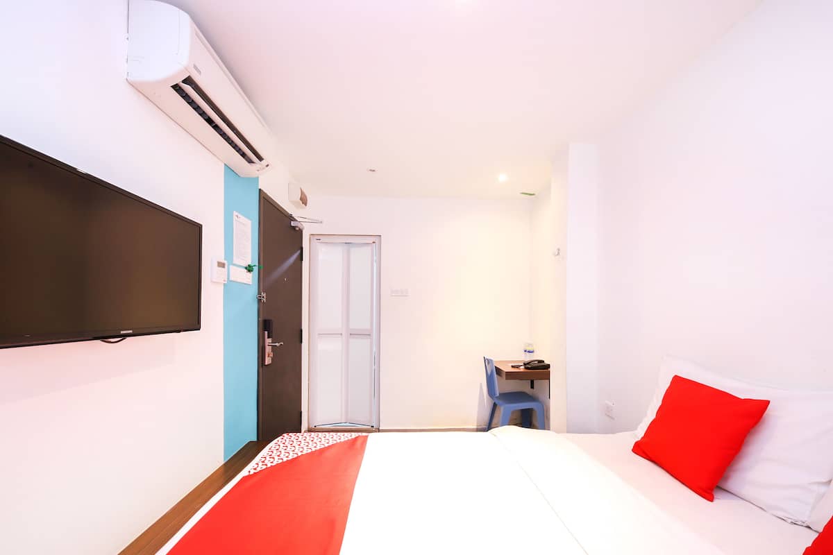 A hotel room with a bed, a desk, a chair, a TV, and an air conditioning unit.