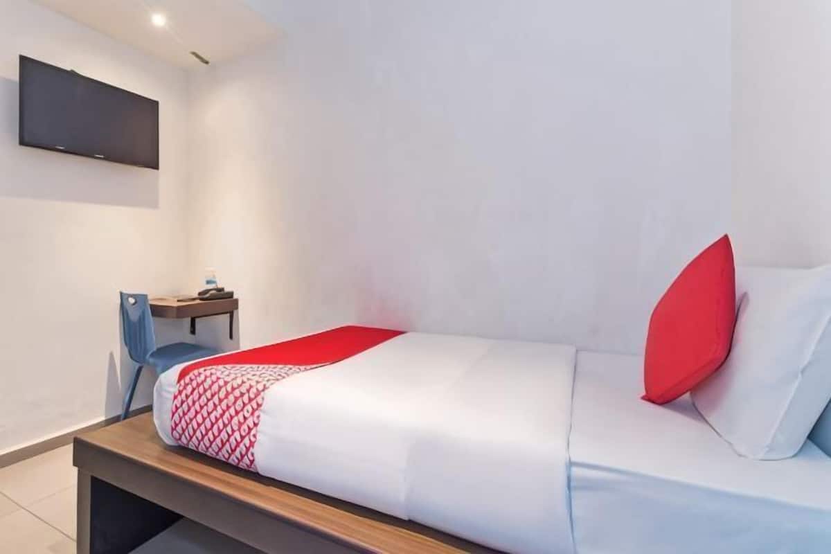 A single bed with a red pillow, a bedside table, a chair, and a flat-screen TV mounted on the wall.