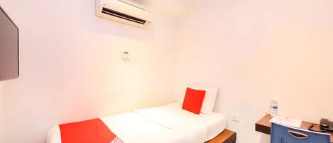 Habitación individual con una cama blanca, manta con estampado rojo y blanco, un banco de madera, un escritorio con una silla y un equipo de aire acondicionado en la pared.