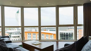 Een moderne hotelkamer met grote ramen van vloer tot plafond, een centraal gelegen zithoek met een koffietafeltje, en uitzicht op de stad en een opvallend gebouw.