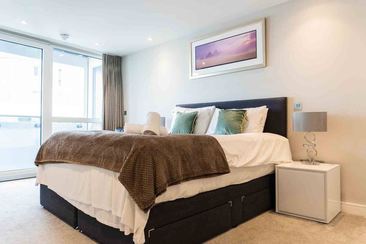 Une chambre moderne avec un grand lit, une table de chevet avec une lampe et une vue sur l’extérieur grâce à une grande fenêtre.