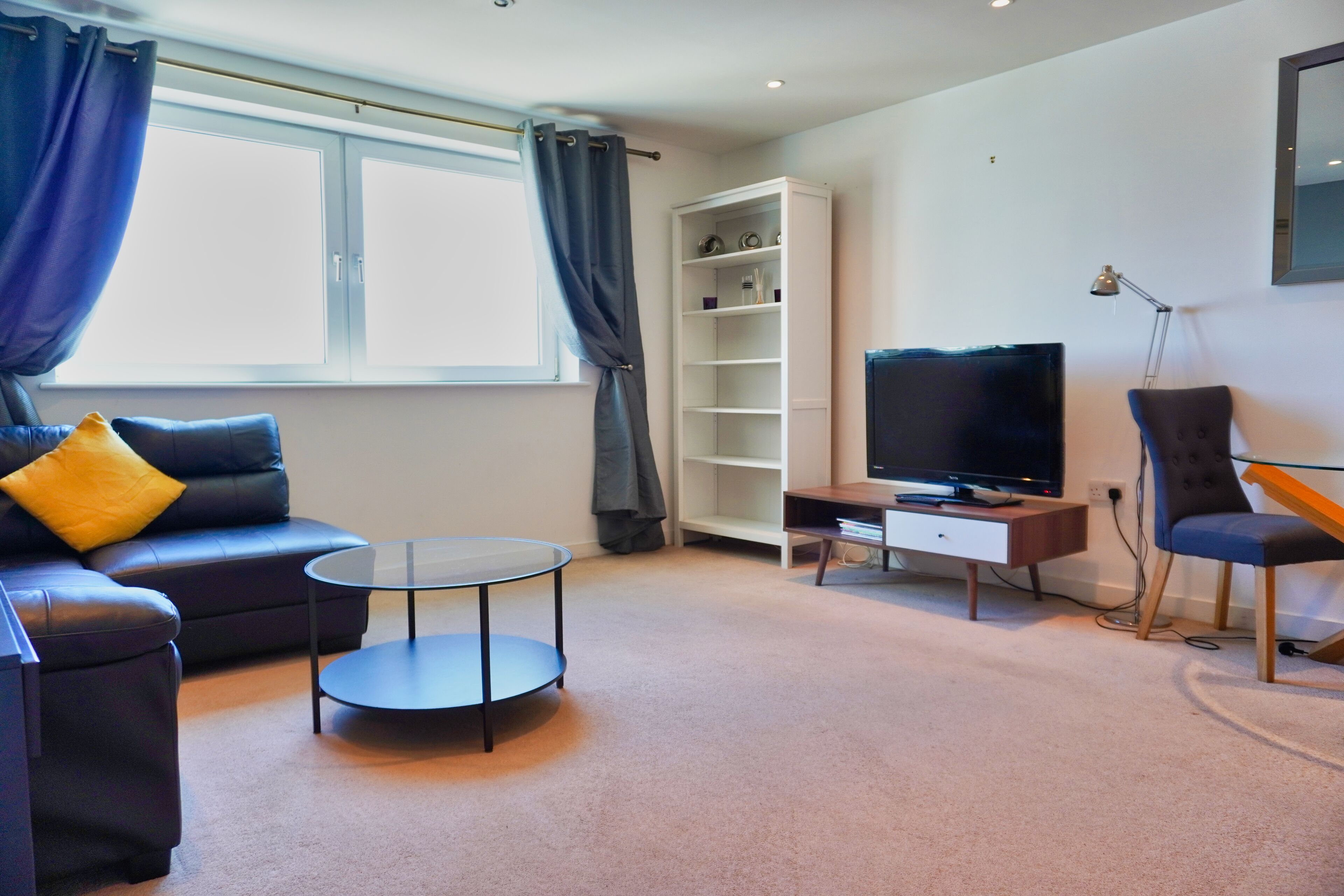 A modern living room with a sofa, a glass coffee table, a TV, a bookshelf, and a lamp.