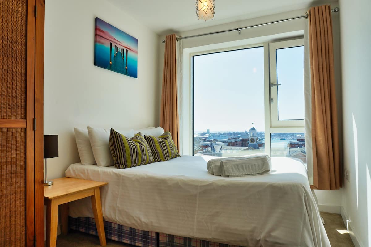 Une chambre d’hôtel avec un lit, une table de chevet, une lampe, un tableau et une vue sur le paysage urbain, visible à travers une grande fenêtre.