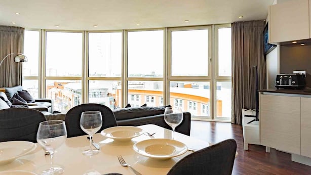 Een eethoek met een tafel voor twee personen, grote ramen, een bank en een keuken met een koffieapparaat.