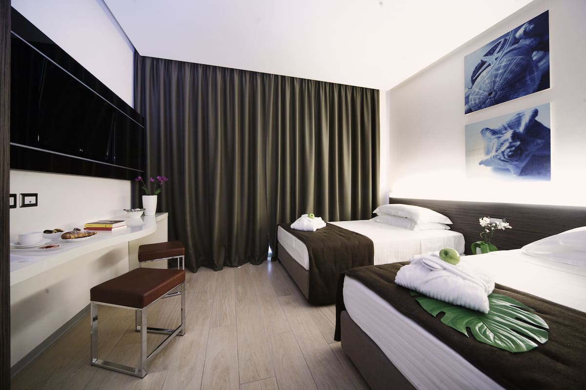 A hotel room with two beds, a desk with a chair, a flat-screen TV, and a large window with curtains.