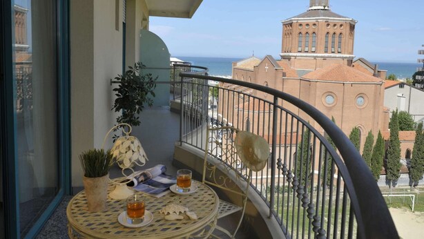 A balcony with a round table and chairs, a potted plant, and a view of a church and the sea. - Bellariva Feeling Hotel (Rimini)