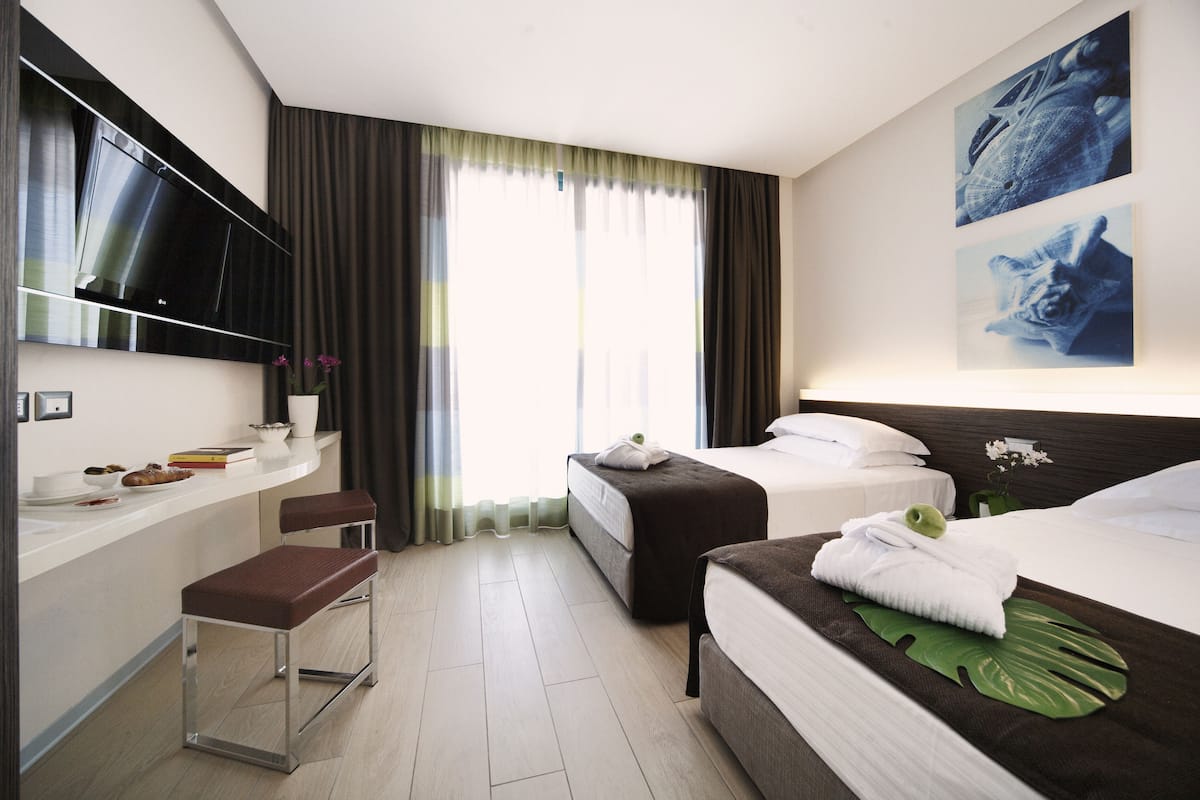A hotel room with two beds, a TV mounted on the wall, a small seating area, and a window with curtains.