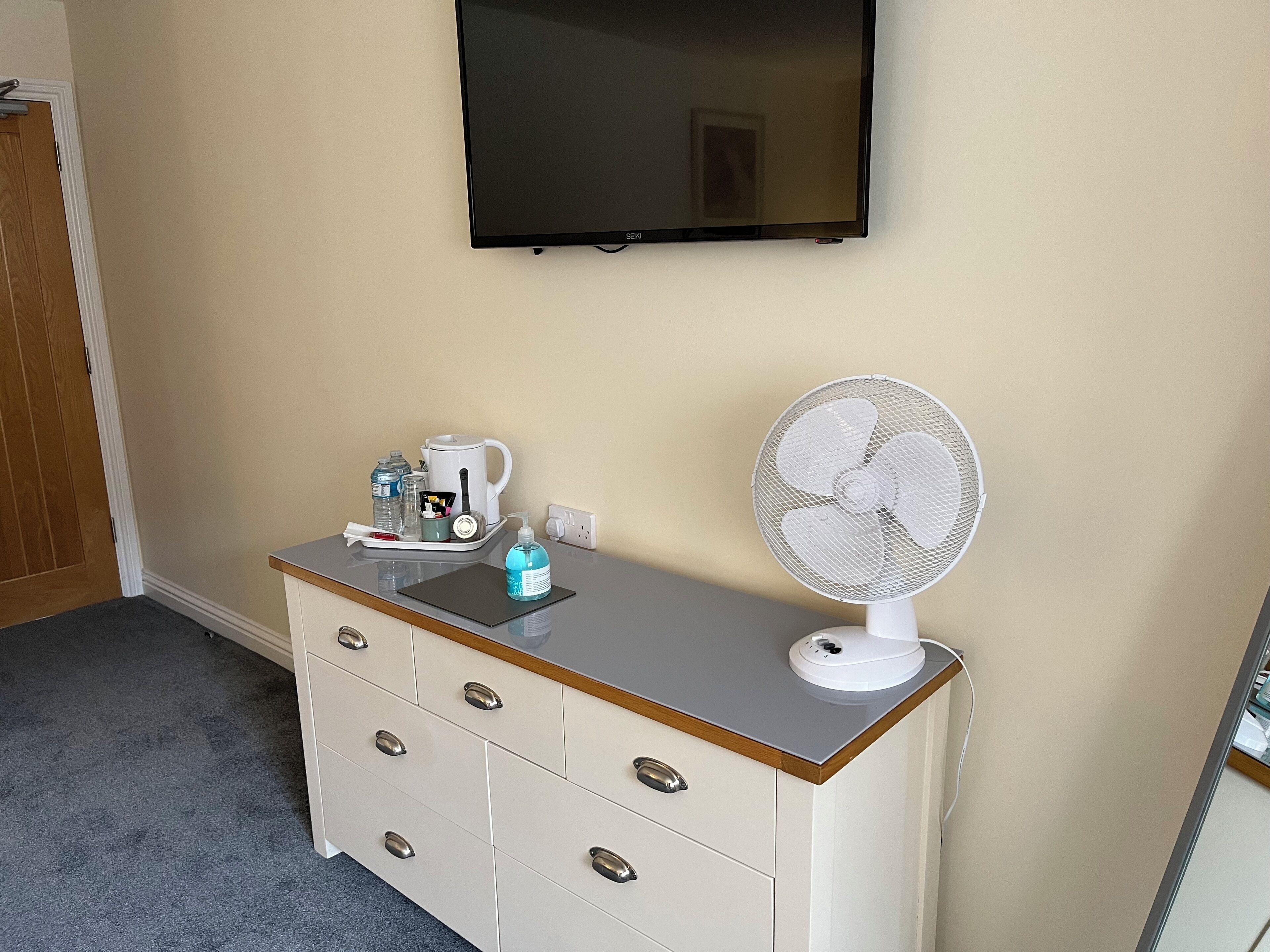Een kamer met een beige wand, een flatscreen-tv boven een witte dressoir met lades, een kleine witte ventilator erop en een koffiezetapparaat op het dressoir.