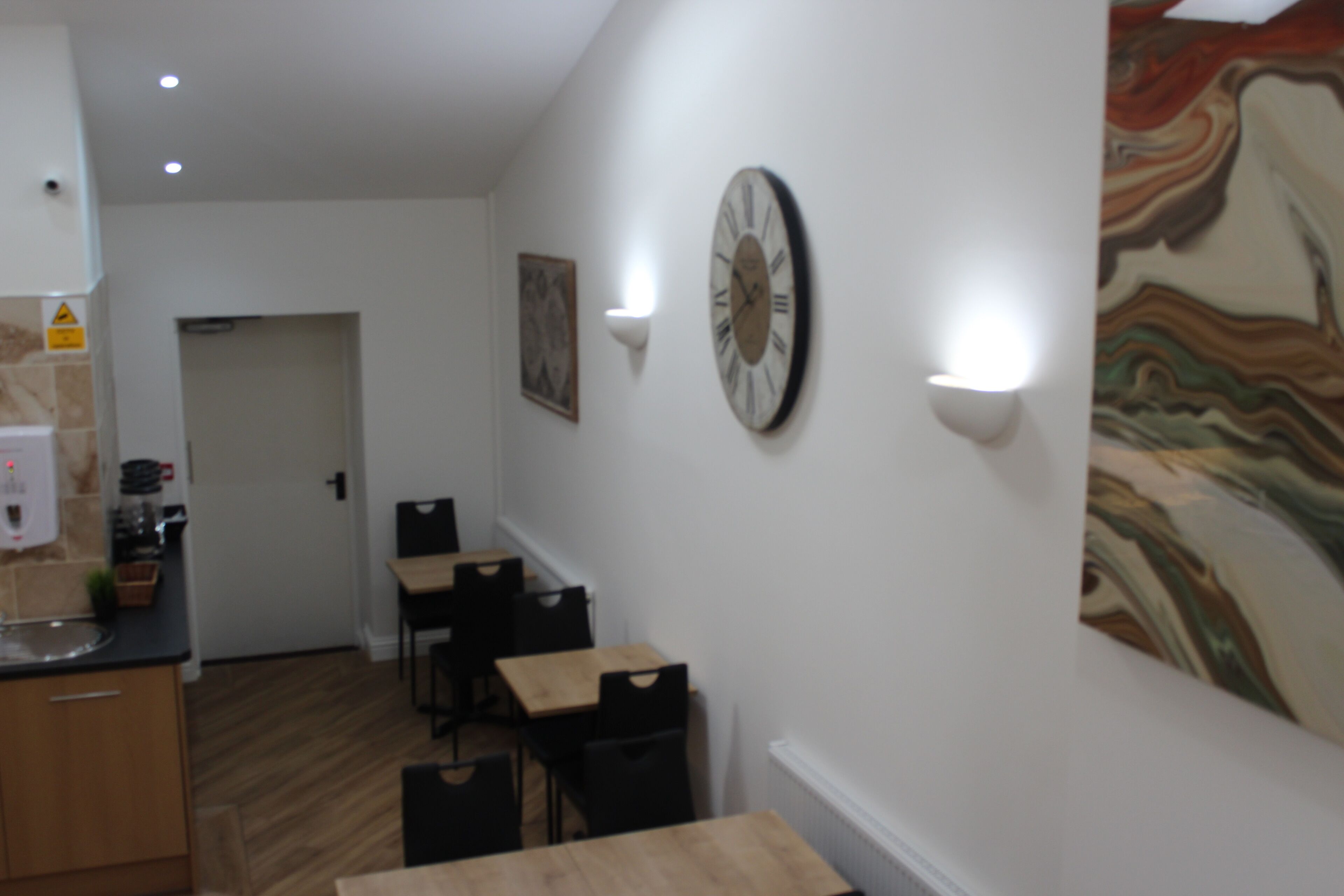 A hallway with a kitchen area on the left, a dining area with tables and chairs, and a large abstract painting on the right wall.
